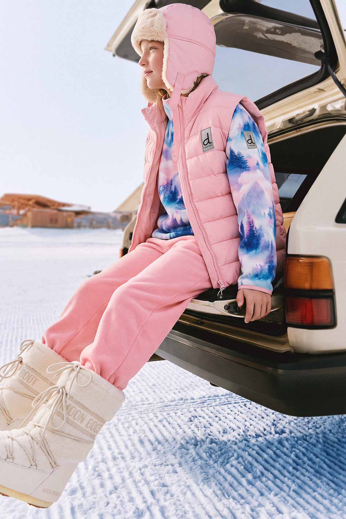 Child sitting on a car tailgate in the snow wearing the light pink Deux par Deux lined chapka and a pink puffer vest