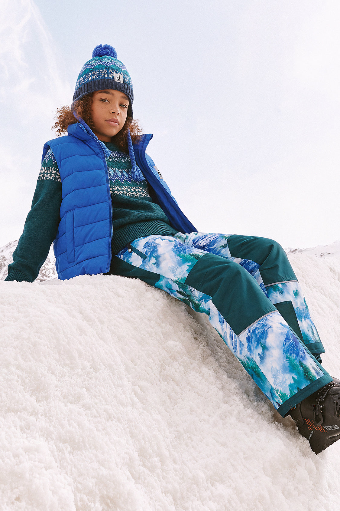Child wearing the Deux par Deux Peruvian Fair Isle knit hat in dark turquoise, seated in the snow