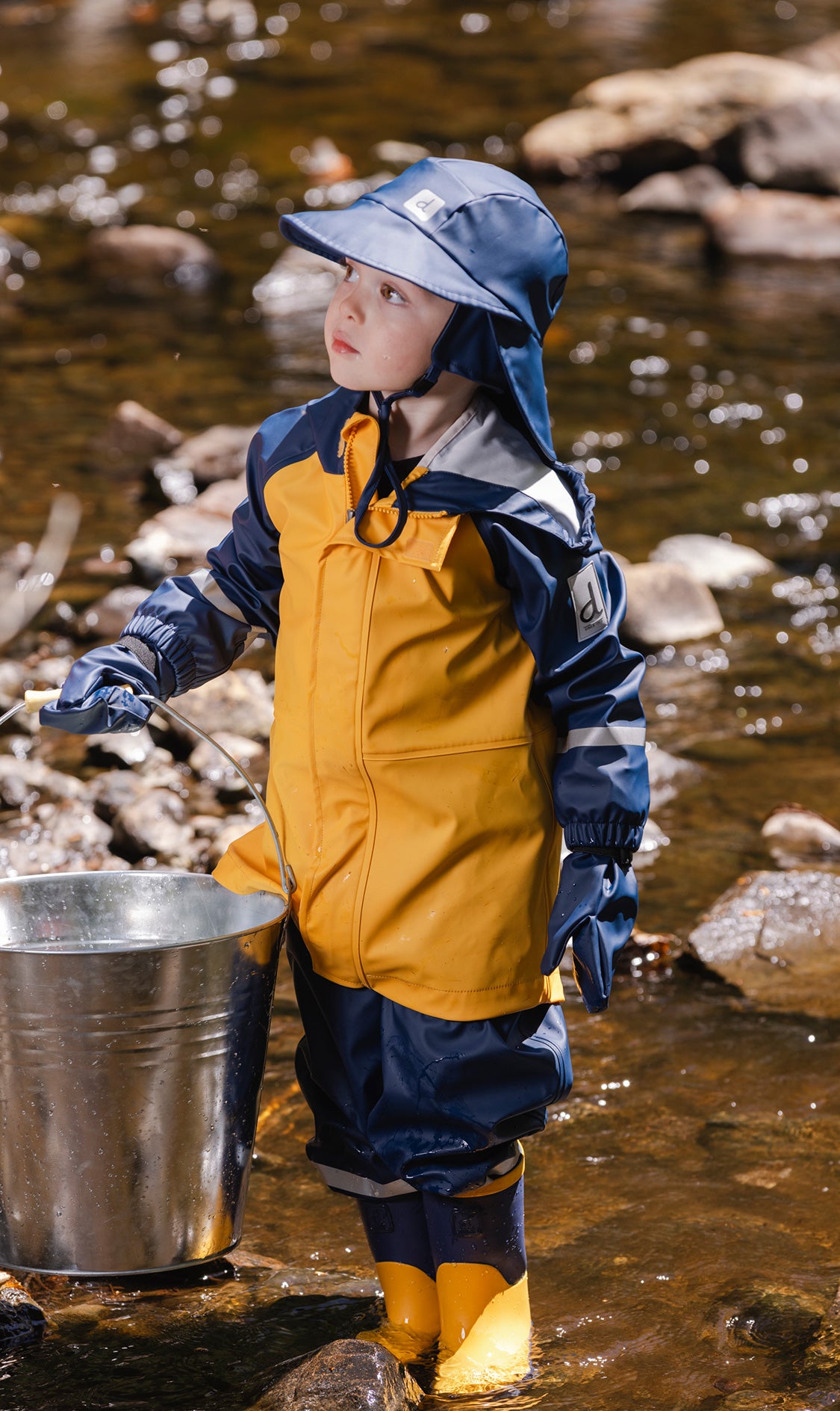 Child in a yellow and navy rain jacket, hat and dark blue rain pants carrying a bucket in a stream