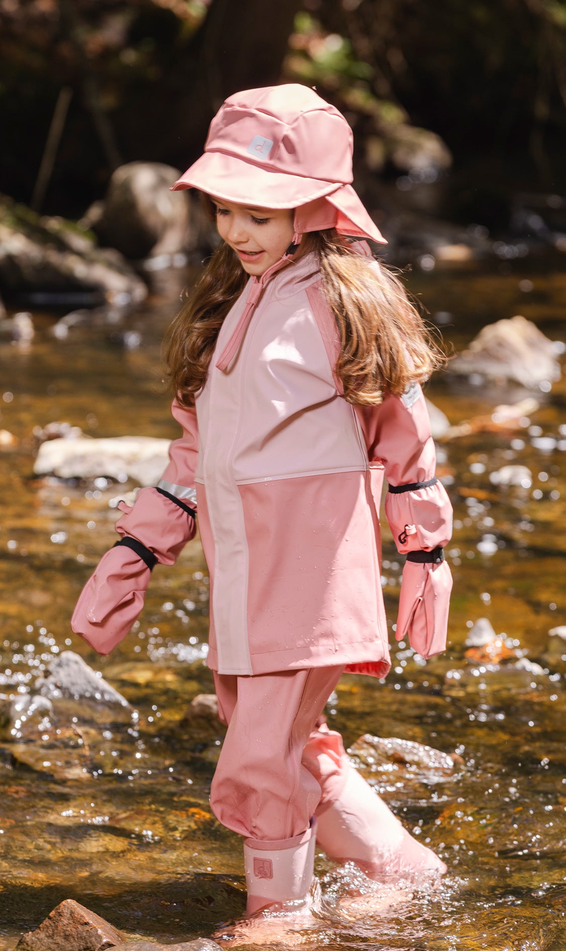 Child wearing the pink Deux par Deux rain overalls with a matching pink rain jacket and hat in a stream