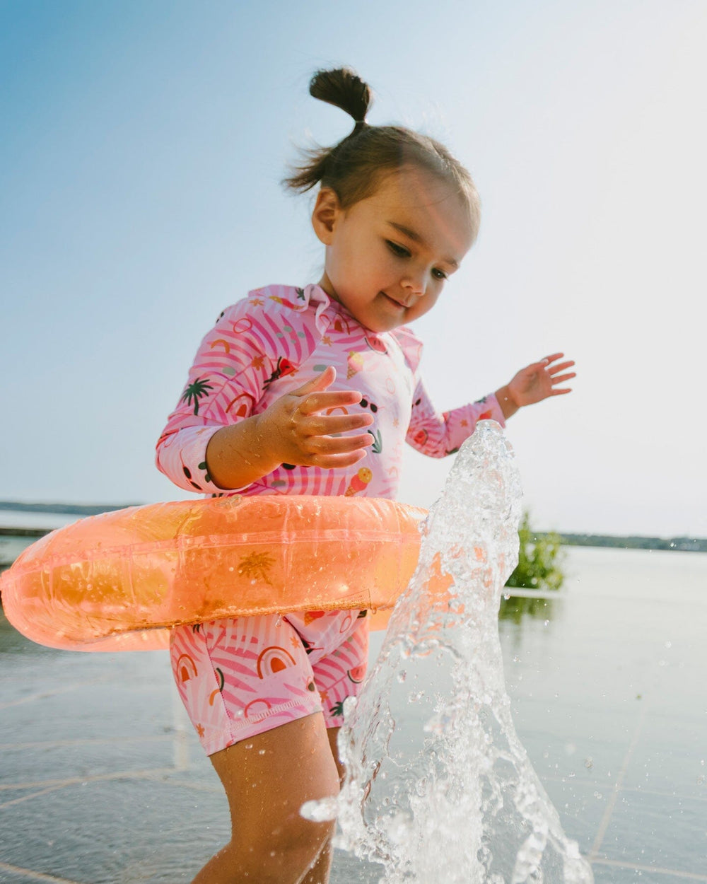 Toddler girl splashing at the beach wearing pink beach print one-piece swimsuit side view
