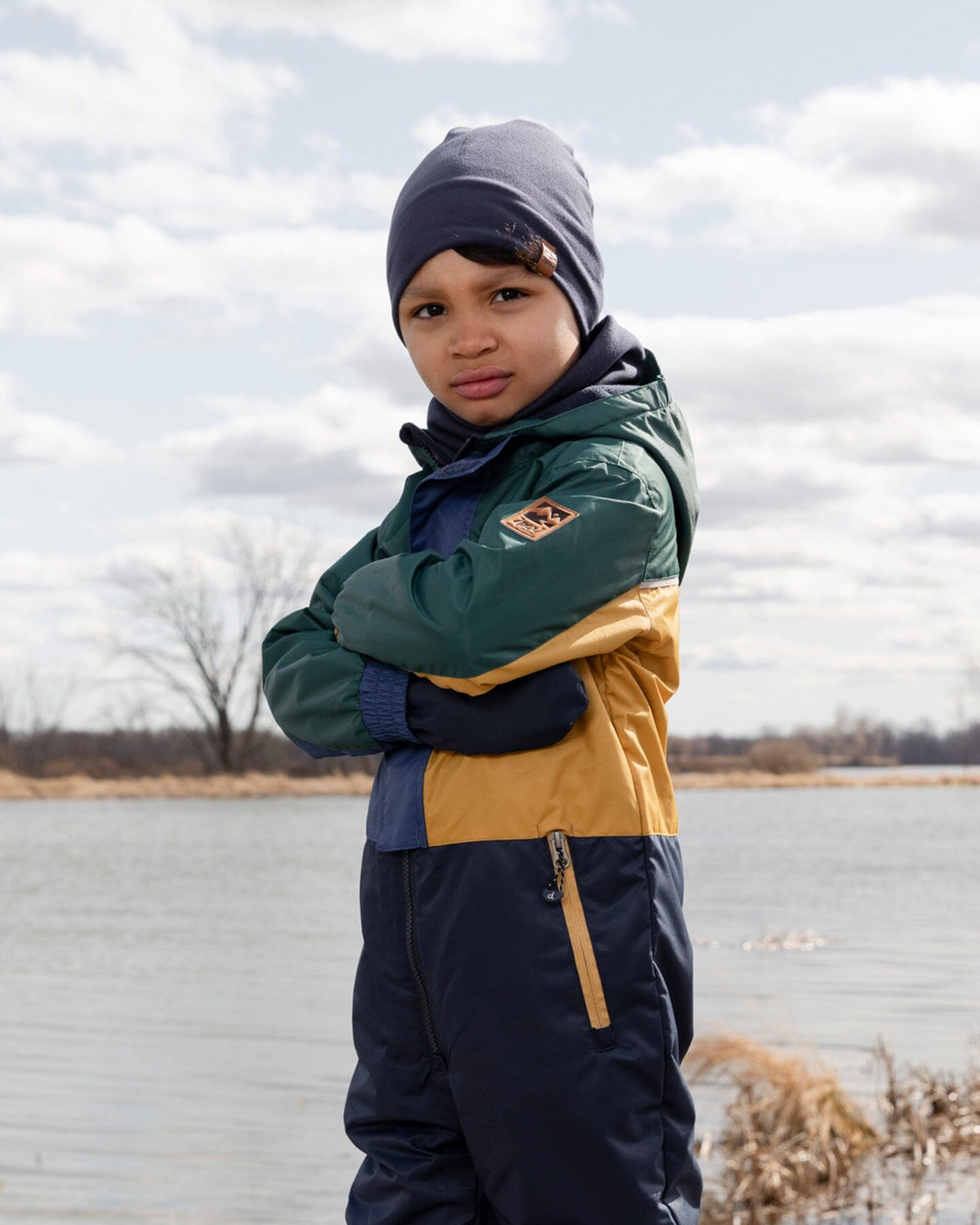 Child standing by a lake in a green, mustard and navy outfit with a navy hat