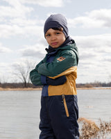 Child standing by a lake in a green, mustard and navy outfit with a navy hat