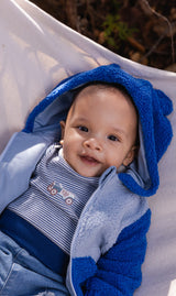 Baby wearing the Deux par Deux two-tone blue sherpa hooded jacket with bear ears, lying down outdoors