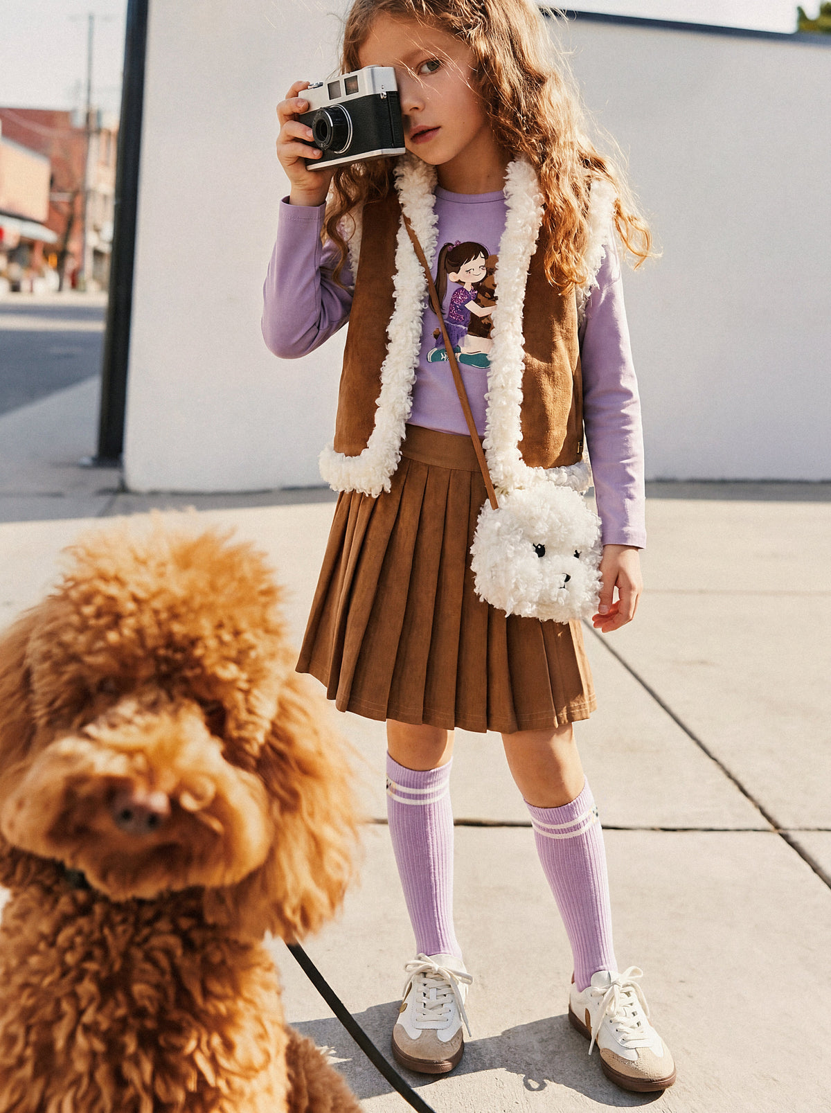 Child wearing the reversible vest, brown side out, with a pleated skirt and a fluffy bag while holding a camera outdoors
