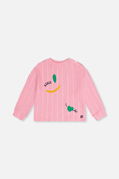 Deux par Deux pink knit top laid flat, front view with a winking face motif and a small green heart