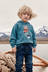 Child wearing the teal snowboarding bear hoodie in a wooden boat with mountains behind