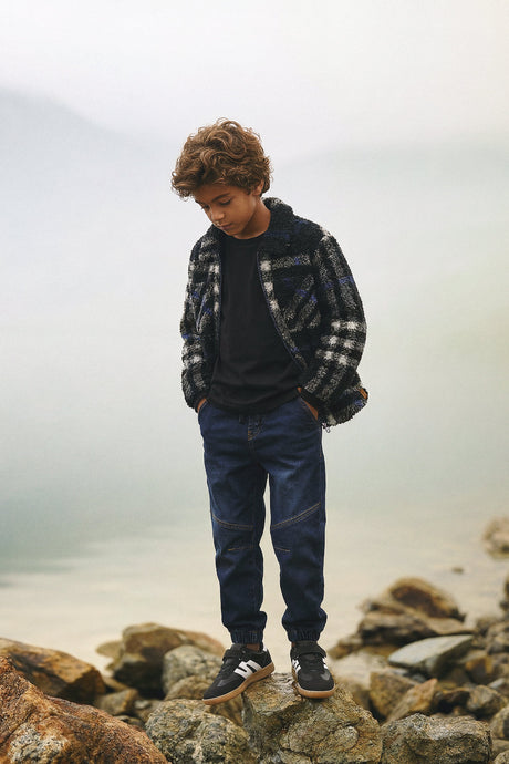 Child wearing the black plaid fleece overshirt with jeans and sneakers on rocks by a misty lake