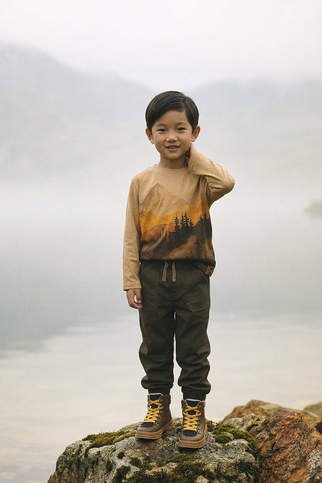Child wearing the beige mountain graphic tee while standing on a lakeside rock