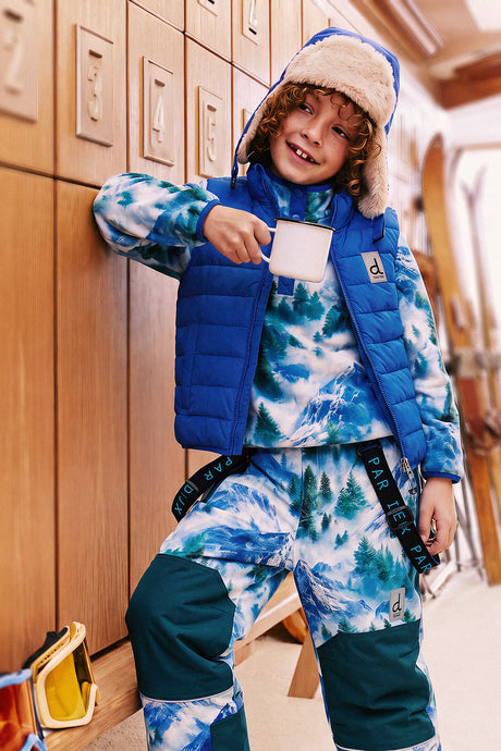 Child leaning on a wooden locker wall in the blue mountain print fleece pullover under a blue vest, holding a mug