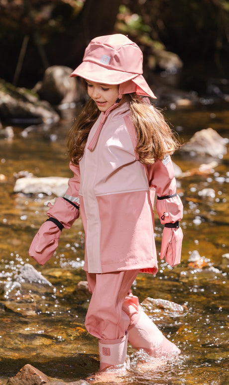 Child wearing the pink Deux par Deux rain overalls with a matching pink rain jacket and hat in a stream