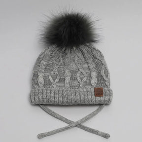 Greymix cable-knit CALIKIDS baby hat with dark faux-fur pompom and chin ties, front view