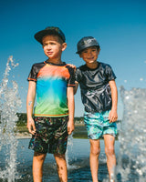 Two boys wearing gradient kids rashguard and matching swim trunks standing on the beach