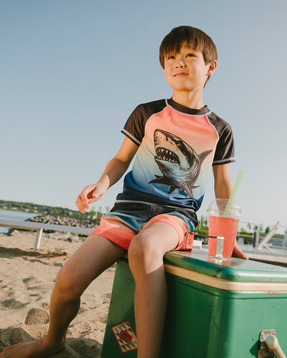 Boy sitting near the water wearing blue/orange gradient swim shorts and rashguard