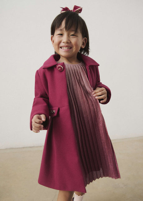 Child wearing the raspberry collared coat over a pleated dress
